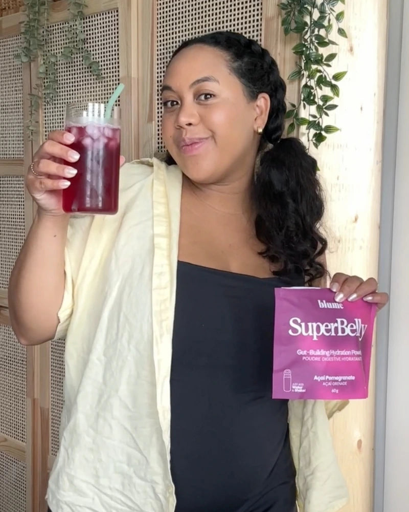 A person smiles while holding a drink and a packet labeled SuperBelly in a kitchen. They wear a black top and light cardigan. There is a green plant in the background.